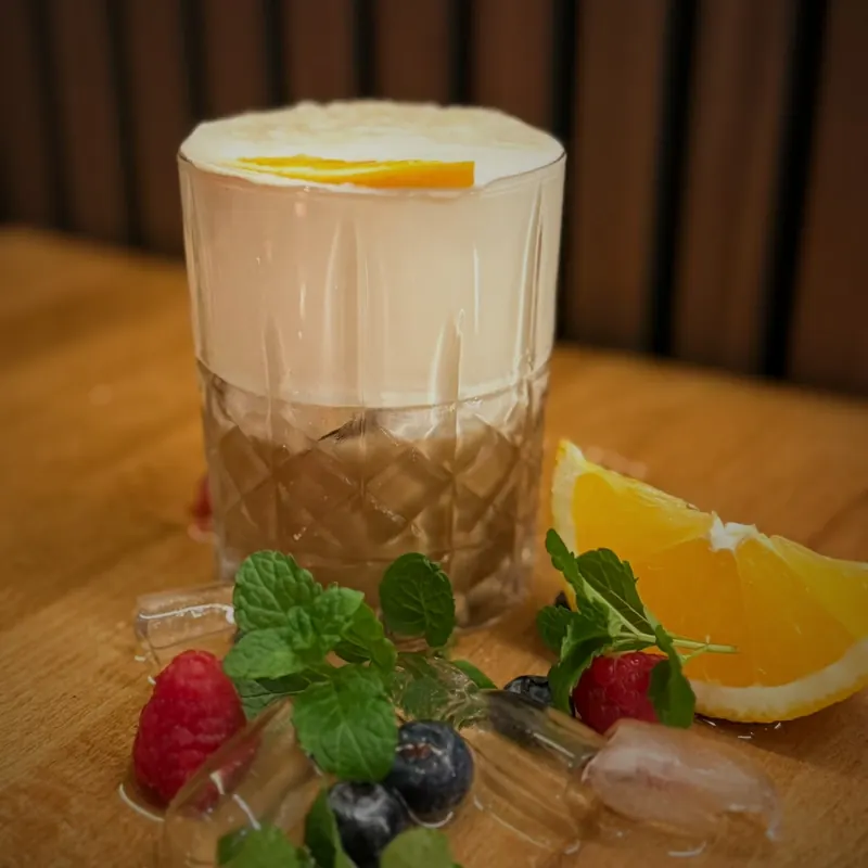 Ein kurzes geschliffenes Glas mit einem hellen Getränk unter einer dicken weißen Schaumkrone, garniert mit einer Orangenscheibe, auf einem Holztisch zwischen Himbeeren, Heidelbeeren, Minze, Eiswürfeln und einem Orangenstück.