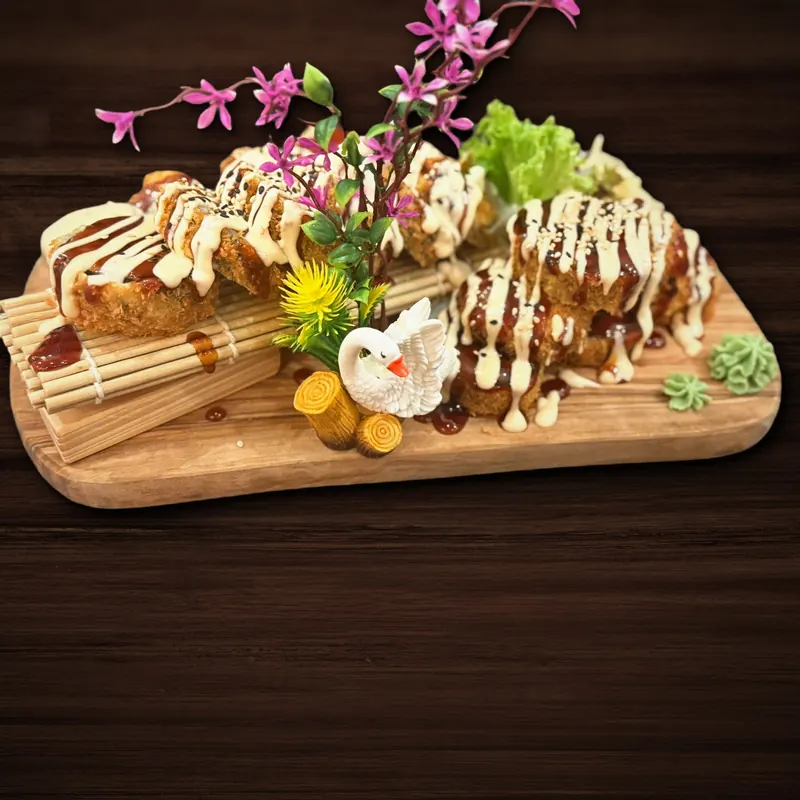 Panierte, frittierte Sushi-Stücke mit Streifen aus dunkler und heller Soße und Sesam auf einem Holzbrett, daneben Wasabi, ein Salatblatt, Dekoblumen und eine kleine Schwanenfigur.