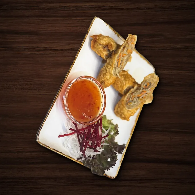 Frittierte Rollen, teils aufgeschnitten mit sichtbarer Füllung aus Karottenstreifen und Glasnudeln, daneben ein Glasschälchen mit orange-roter Chilisoße, Rettich- und Rote-Bete-Streifen und Salat auf einem weißen Teller.