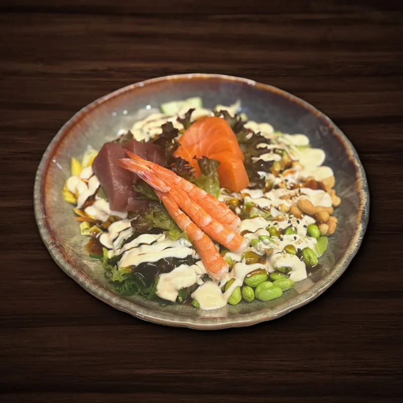 Bowl mit rohen Thunfisch- und Lachsscheiben, zwei Garnelen, Salat, Algensalat, Edamame, Mango und Nüssen, beträufelt mit heller und dunkler Soße.