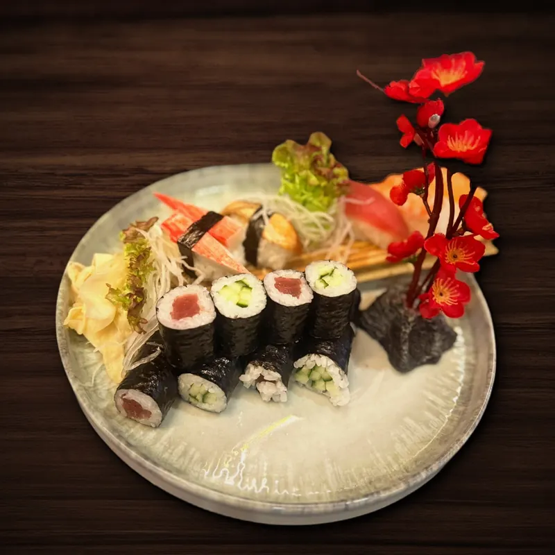 Runder Teller mit Maki-Röllchen mit Thunfisch- oder Gurkenfüllung, dahinter Nigiri mit Surimi und Thunfisch, dazu eingelegter Ingwer, Salatblätter und ein Dekozweig mit roten Blüten.