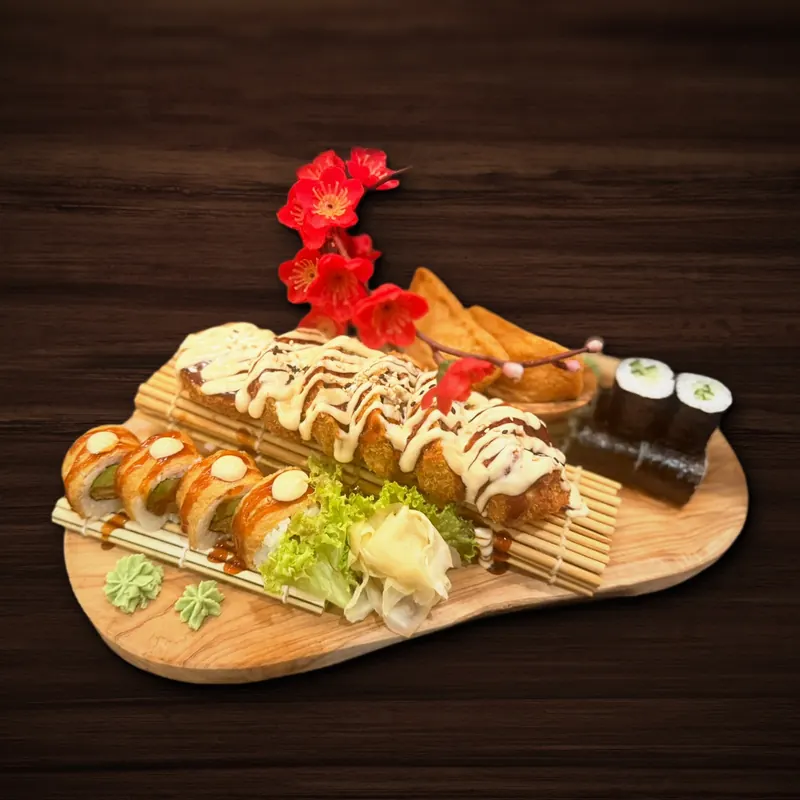 Holzbrett mit einer ganzen, panierten Sushi-Rolle unter heller und dunkler Soße, davor Rollenstücke mit Avocado in goldgelber Hülle, dahinter frittierte Teigtaschen und Gurken-Maki, dazu Wasabi, Ingwer, Salat und ein Dekozweig mit roten Blüten.